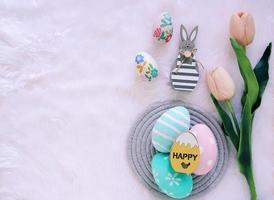 Happy Easter concept with wooden bunny and colorful easter eggs on white fur background and pink tulips. Top view with copy space photo