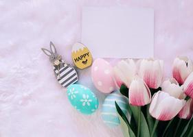 Happy Easter concept with wooden bunny and colorful easter eggs on white fur background and pink tulips. Top view with copy space photo