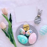 Happy Easter concept with wooden bunny and colorful easter eggs on white fur background and pink tulips. Top view photo