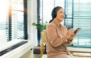 Grandmother listen old music in headphone near piano. Elderly Senior Asian woman enjoy relax from song wireless headset which play oldies songs as think old day and remember cool person, copy space photo