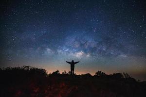 Landscape with milky way galaxy, Starry night sky with stars and silhouette of a standing sporty man with raised up arms on high mountain. photo