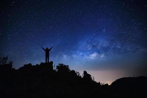paisaje con galaxia de la vía láctea, cielo nocturno estrellado con estrellas y silueta de un hombre deportivo de pie con los brazos levantados en la alta montaña.