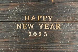 Happy New year 2023 celebration. Wooden text on wooden background with scattered golden confetti. Flat lay. photo