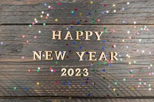 Happy new year 2023 quotation on wooden background with scattered confetti of multicolored stars. Festive greeting card photo