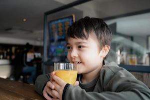 Side view Portrait Young kid drinking fresh orange juice for breakfast in cafe, Happy child boy drinking glass of fruit juice while waiting for food in restaurant, Healthy food lifestyle concept photo