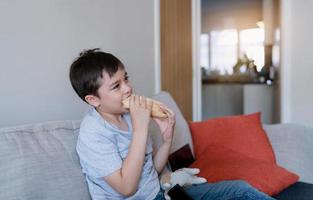 Hungry kid boy eating homemade bread sandwiches with mixed vegetables, Child siting on sofa having lunch while watching TV, Cute boy relaxing at home eating take way food on Summer school break. photo