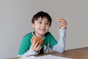 Healthy young boy eating burger. A child holding cheeseburger and looking at chips on his hand with smiling face, Happy Kid having fast food hamburger for snack. Favorite children's food. photo