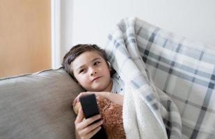 Kid sitting on sofa watching TV, A boy holding remote control and looking up with smiling face, Positive child lying on sofa enjoy watching cartoon on TV, Kid relaxing at home on weekend photo