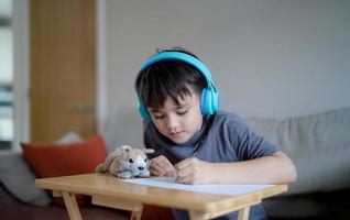Happy kid wearing some headphones and listening to music while drawing, sketching on paper, Indoor portrait by Cute boy enjoy a creative activity at home on a weekend. A Child is doing homework photo