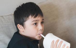 Portrait happy Kid drinking water or juice from paper cup, Child boy sitting on sofa drinking cold soda while watching TV relaxing at home on Spring or Summer photo