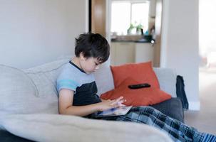 retrato de un niño jugando en una tableta sentado en un sofá, un niño leyendo o haciendo la tarea en línea en Internet en casa, un niño viendo dibujos animados en línea en una almohadilla digital. educación en el hogar, concepto de regreso a la escuela foto
