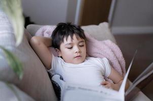 Happy School kid lying on sofa reading a book with morning light shining from window. Child boy reading story relaxing at home on weekend, Positive children concept photo