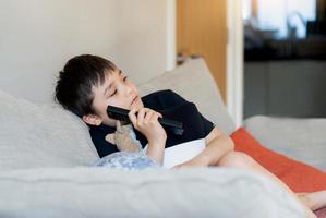 Kid sitting on sofa watching TV, A boy holding remote control and looking with deep in thought, Positive child lying on sofa enjoy watching cartoon on TV, Kid relaxing at home on weekend photo