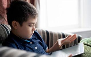 Cinematic portrait young boy playing game on tablet sitting on sofa with light shining from window, Kid playing games online on internet at home, Child talking video call with friends at home photo