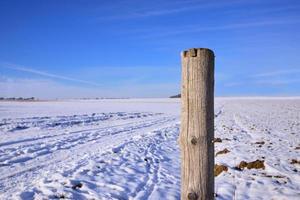 A weathered fence post stands in a wide winter landscape in Bavaria, against snow and a blue sky photo