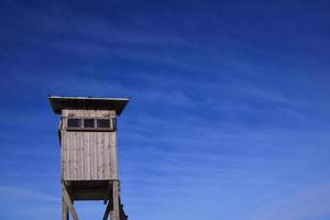 A weathered wooden stand for hunters stands on wooden posts and has a narrow window. The high stand is at the bottom of the picture against a blue sky with clouds in Bavaria