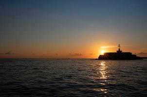 The sun goes down at the sea. The sky glows blue and orange. The light is reflected on the waves. The shadow of a signal tower is in front of the sun at the side of the picture. photo