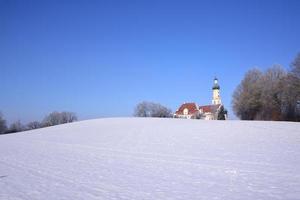 en invierno, la histórica iglesia de peregrinación de biberbach se alza sobre una colina cubierta de nieve contra un cielo azul foto