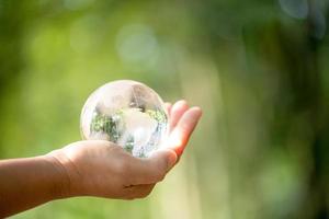 mano humana sosteniendo globo planeta cristal en bosque verde con luces de naturaleza bokeh. día Mundial del Medio Ambiente. concepto para la conservación del medio ambiente, proteger la ecología de la tierra y la vida ecológica. foto