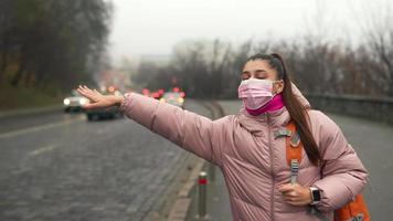 Young woman in pink mask and coat hails for car ride on the side of a busy road video