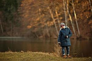 Boy in jacket and sunglasses stand in sunny spring park against river. photo