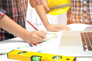 Asian engineer and partner team learn and talk with house plan project blueprint, graph and yellow helmet and engineering construction tools on table work. photo