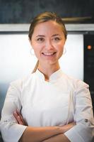 Portrait of confident and smiling young woman chef dressed in white uniform, professional kitchen are on background photo