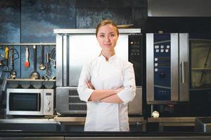 retrato de una joven chef confiada y sonriente vestida con uniforme blanco, la cocina profesional está en segundo plano foto