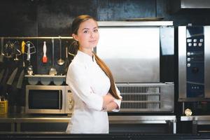 Portrait of confident and smiling young woman chef dressed in white uniform, professional kitchen are on background photo