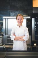 Portrait of confident and smiling young woman chef dressed in white uniform, professional kitchen are on background photo