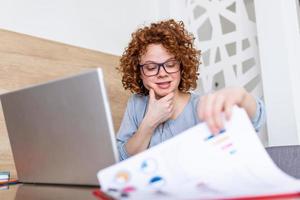 Serious focused businesswoman with laptop holding papers preparing report analyzing work results, female executive doing paperwork at workplace using computer online software for data analysis photo