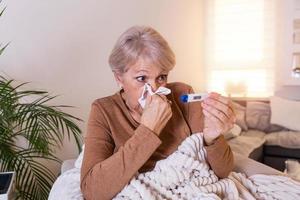Sickness, seasonal virus problem concept. Senior woman being sick having flu lying on sofa looking at temperature on thermometer. Sick older lady lying in bed with high fever. Cold flu and migraine. photo