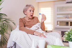 enfermedad, concepto de problema de virus estacional. anciana enferma con gripe tendida en el sofá mirando la temperatura en el termómetro. anciana enferma acostada en la cama con fiebre alta. gripe fría y migraña. foto