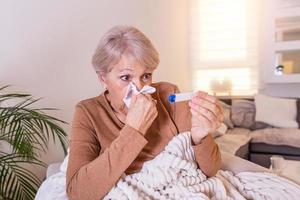 Sickness, seasonal virus problem concept. Senior woman being sick having flu lying on sofa looking at temperature on thermometer. Sick older lady lying in bed with high fever. Cold flu and migraine. photo