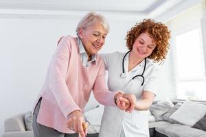 Young carer supporting senior disabled woman with walking stick. Portrait of happy female caregiver and senior woman walking together at home. Professional caregiver taking care of elderly woman. photo