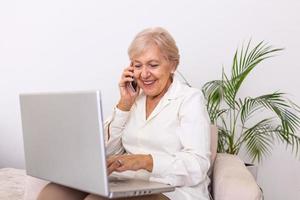 Elderly woman working on laptop computer, smiling, talking on the phone. Senior woman using laptop. Elderly woman sitting at home, using laptop computer and talking on her mobile phone, smiling. photo