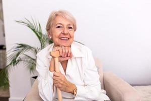 retrato de una hermosa anciana con cabello blanco y bastón. retrato de una anciana sentada en un sofá en casa. Sonriente mujer madura de pelo gris de mediana edad mirando a la cámara foto