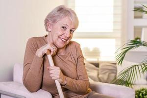 Portrait of beautiful senior woman with white hair and walking stick. Portrait of senior woman sitting on sofa at home. Smiling middle aged mature grey haired woman looking at camera photo