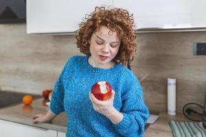 Portrait of a lovely smiling pretty girl biting an apple in her kitchen. Young woman enjoying her red organic apple fruit. Daily intake of vitamins with fruits, Diet and healthy eating photo