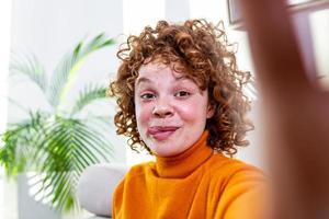 Crazy young female with curly hair showing tongue while taking selfie. Close up photo of beautiful a red hair girl taking selfies hand arm holding camera broadcasting blog wear casual sweater pullover