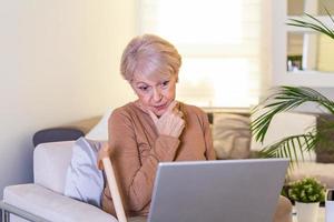 Senior woman working on home finance. Mature woman checking bills at home. Older woman checking pension approval certificate with laptop at home. photo