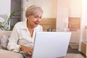 una anciana usando una computadora portátil. retrato de una hermosa mujer mayor que trabaja con una computadora portátil en el interior. mujer mayor sentada en un sofá con una laptop en casa foto