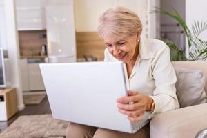 mujer madura feliz que tiene una videollamada a través de una laptop en casa. anciana de cabello gris ondeando la mano frente a la computadora portátil mientras realiza una videollamada con los miembros de su familia. foto