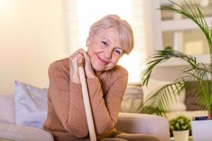 Pleasant thoughtful aged woman seating with a walking stick. Retired woman with her wooden walking stick at home. Happy senior woman relaxing at home holding cane and looking at camera. photo