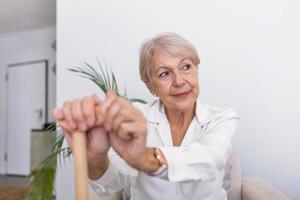 Elder lady sitting on the couch with wooden walking stick and smiling. Happy elderly woman relaxing on sofa and holding walking stick. Copy space. Senior woman looking thoughtful in a retirement home photo