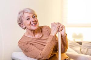 Elder lady sitting on the couch with wooden walking stick and smiling. Happy elderly woman relaxing on sofa and holding walking stick. Copy space. Senior woman looking thoughtful in a retirement home photo