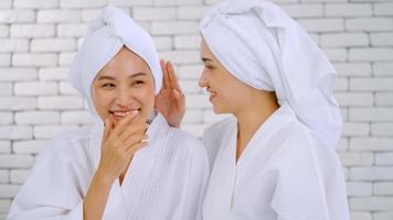 dos chicas asiáticas en albornoces blancos con toallas en la cabeza hablando en la sala de estar. foto