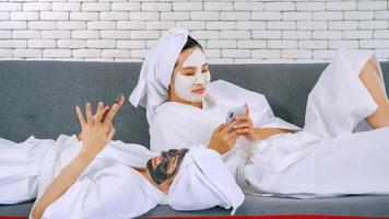 Two happy Asian girls in white bathrobes with facial mask using smart phone on sofa. photo