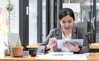 bella y sonriente mujer de negocios asiática con anteojos sosteniendo una pluma trabajando con una tableta en la oficina. foto