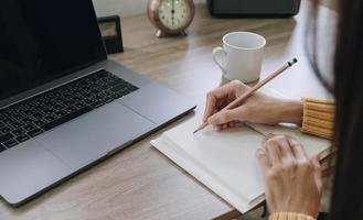 woman writing taking notes while sitting in front her computer laptop at the wooden working table over living room photo